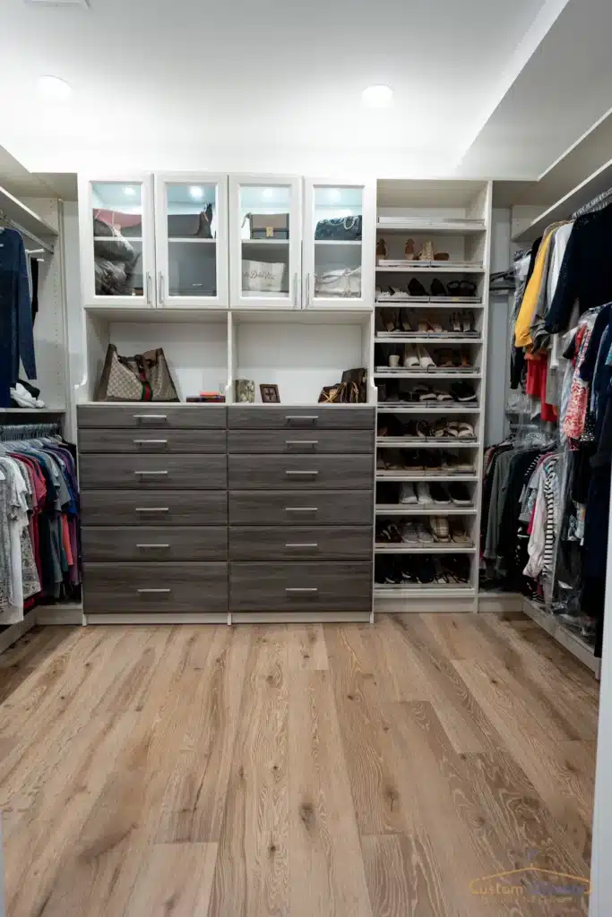 custom walk in closet with brown drawers and white upper shelves hidden by glass doors. and floor mounted shoe closet is next to it, on the side features some hanging space